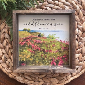 May include: Square wooden sign with a landscape print of wildflowers and the text "Consider how the wildflowers grow" from Luke 12:27. The sign has a dark brown frame and is set on a woven mat.