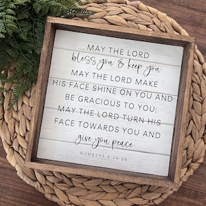 May include: A white wooden sign with a brown frame, featuring a religious quote: "May the Lord bless you & keep you. May the Lord make His face shine on you and be gracious to you; May the Lord turn His face towards you and give you peace." The quote is attributed to Numbers 6:24-26.