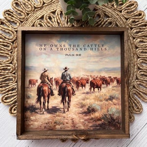 May include: A framed rustic wooden sign featuring a Western landscape painting with cowboys herding cattle. The text "HE OWNS THE CATTLE ON A THOUSAND HILLS" and "PSALM 50:10" are printed on the artwork. The sign is displayed on a woven placemat.