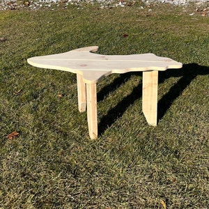 May include: A wooden coffee table shaped like the state of Michigan. The table is made of natural wood and has a rustic finish. It is sitting on a grassy lawn.