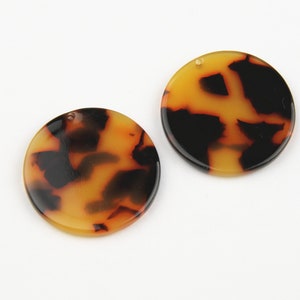 Puede incluir: Dos colgantes redondos con un patrón de carey, compuestos por tonos negros y ámbar. Cada colgante tiene un pequeño agujero para ensartar. La superficie brillante resalta el diseño moteado. Ideal para la creación de joyas.