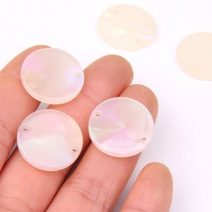 Puede incluir: Cuatro botones redondos de concha blanca iridiscente con dos agujeros cada uno. Los botones están en una mano y una superficie blanca.