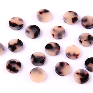May include: A collection of 15 round, flat, white and brown patterned resin buttons. The buttons have a mottled, tortoiseshell-like pattern.