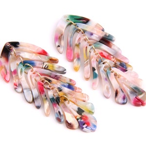 May include: A pair of colorful, multi-layered, acrylic earrings. Each earring is shaped like a feather and features a variety of colors, including pink, green, blue, and yellow.
