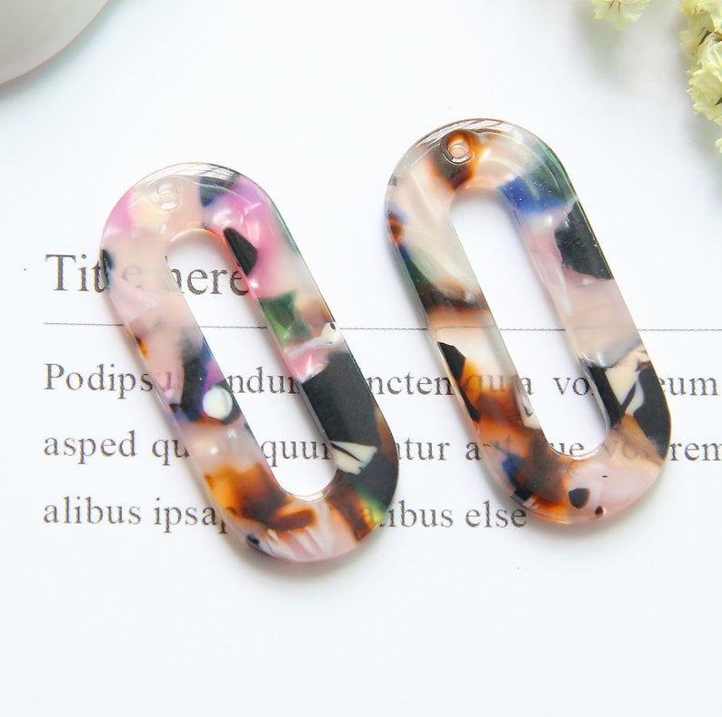 May include: Two oval-shaped, colorful, plastic, resin, or acrylic,  earring blanks with a hole at the top. The blanks have a mottled pattern of pink, brown, black, and white.