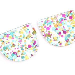Puede incluir: Dos formas de medio círculo de resina transparente con confeti dorado, rosa, azul y turquesa incrustados.