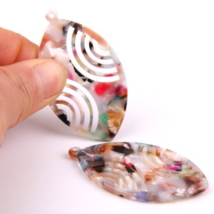 Puede incluir: Dos pendientes ovalados de acrílico de colores con un patrón geométrico blanco. Los pendientes tienen un agujero en la parte superior para colgar.