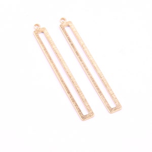 May include: Two gold-colored metal rectangular pendant blanks with a textured surface. The blanks have a loop at the top for attaching to a chain or cord.