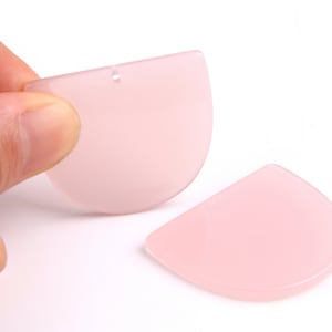 Puede incluir: Dos colgantes semicirculares de color rosa pálido. Uno está sostenido por una mano, mostrando un pequeño agujero en la parte superior para ensartar. El otro colgante está plano. Los colgantes tienen un aspecto liso y translúcido.