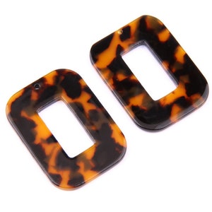 Può includere: Due orecchini rettangolari in plastica marrone e nera con un foro nella parte superiore per appenderli. Gli orecchini hanno un motivo a tartaruga.