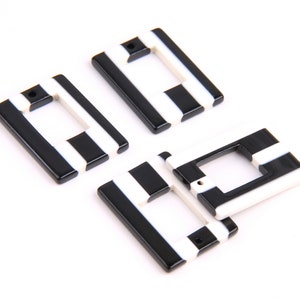 Puede incluir: Cuatro dijes rectangulares de plástico con rayas blancas y negras y un agujero en el centro.