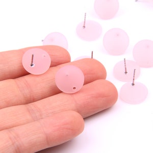 Op de afbeelding: Een close-up van een hand die verschillende kleine, ronde, lichtroze oorbelstuds met zilveren pinnen vasthoudt. De oorbellen zijn verspreid over een wit oppervlak.