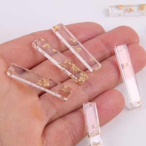 May include: A set of six clear rectangular resin charms with gold leaf accents. The charms are approximately 1 inch long and 1/2 inch wide.