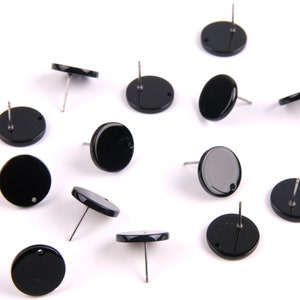 Puede incluir: Una colección de doce pares de pendientes redondos negros con postes plateados. Los pendientes están esparcidos sobre una superficie blanca.