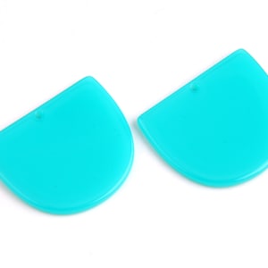 Puede incluir: Dos colgantes de resina acrílica de color azul turquesa en forma de semicírculo con un agujero en la parte superior para colgar.