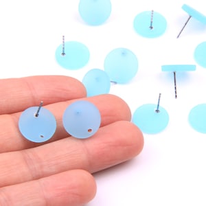 Puede incluir: Primer plano de una mano sosteniendo un par de pendientes redondos de color azul claro. Los pendientes están hechos de vidrio esmerilado y tienen postes de plata.