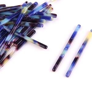 Puede incluir: Un montón de palitos para el pelo de plástico de colores azul, amarillo y morado. Dos palitos están colocados planos sobre una superficie blanca.