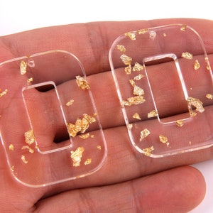 May include: Two clear rectangular resin earrings with gold flakes embedded inside. The earrings have a rectangular hole in the center.