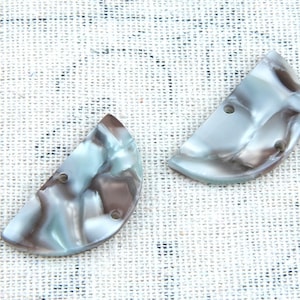 Puede incluir: Dos botones de plástico en forma de semicírculo con un patrón de mármol en tonos marrones, blancos y azules. Cada botón tiene dos agujeros.