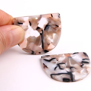 Puede incluir: Dos colgantes de resina en forma de semicírculo con un patrón de mármol en tonos de blanco, negro y rosa.