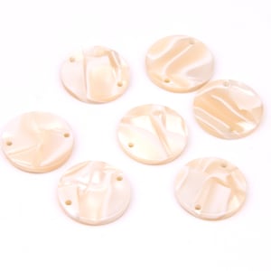 Puede incluir: Siete cuentas redondas de color crema con efecto veteado y dos agujeros cada una. Estas cuentas son adecuadas para la fabricación de joyas y proyectos de artesanía. Las cuentas están dispuestas sobre una superficie blanca.