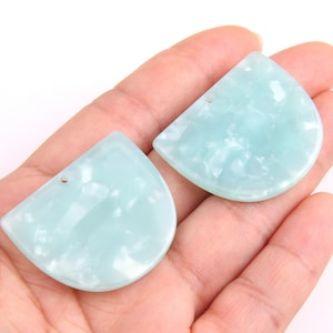 Puede incluir: Dos colgantes semicirculares de color azul claro con efecto veteado. Cada colgante tiene un pequeño agujero cerca de la parte superior para ensartar. Los colgantes se sostienen en una mano sobre un fondo blanco. Adecuado para la fabricación de joyas.