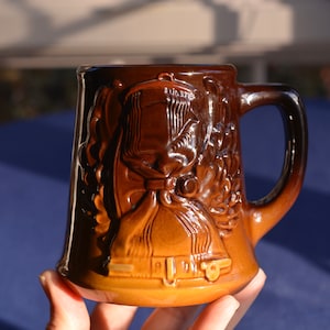 May include: A brown ceramic mug with a raised design of a boot and a floral motif. The mug has a handle and is glazed in a dark brown color.