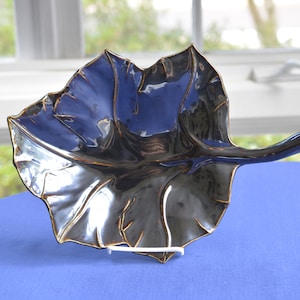 May include: A black ceramic leaf-shaped bowl with a glossy finish. The bowl has a raised, textured design that resembles veins on a leaf. The bowl is on a blue surface.