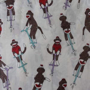 May include: White fabric with a repeating pattern of brown monkeys riding on pogo sticks. The monkeys are wearing red, white, and blue hats and have a sock monkey design.