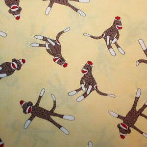May include: A yellow fabric background with a repeating pattern of brown sock monkeys. The monkeys are in various poses, with some standing, some sitting, and some lying down. The monkeys have red noses and mouths, and some have red hats.