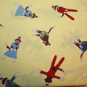 May include: A yellow fabric with a repeating pattern of sock monkeys in various poses. The monkeys are wearing different colored clothing and have different facial expressions.