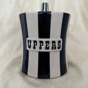 May include: A cylindrical ceramic canister featuring vertical black and white stripes. A central rectangular plaque on the front displays the word UPPERS in bold black lettering. The canister includes a matching lid with a small central handle.