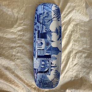 May include: A long, rectangular ceramic serving tray with rounded corners, featuring a blue and white transferware pattern depicting a detailed landscape with a large building, trees, and a boat on a river, finished with a decorative border.