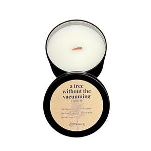 May include: A black metal container holds a white candle with a wooden wick. The lid, also black, features text that reads "a tree without the vacuuming" and "Frasier fir." The candle is handpoured coconut wax and has a 30-hour burn time.