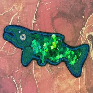 Pode incluir: Um patch verde em forma de peixe com contorno azul e preenchido com lantejoulas verdes. O peixe tem um olho e uma boca detalhados. O patch está em um fundo marmorizado rosa e dourado.