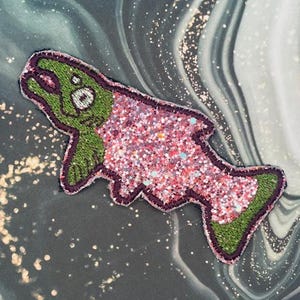 Puede incluir: Parche bordado con forma de pez de dibujos animados. El pez tiene la cabeza y la cola verdes, con un cuerpo rosa y brillante. El parche está delineado con hilo morado oscuro.