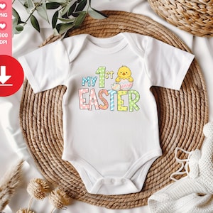 Könnte beinhalten: Weißer Baby-Body mit dem Text "My 1st Easter" in bunten Buchstaben und einem Cartoon-Küken, das aus einem Ei schlüpft. Der Body liegt auf einer gewebten, kreisförmigen Matte mit einer weißen Decke und Grünzeug im Hintergrund.