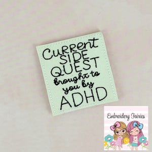 Könnte beinhalten: Ein hellgrüner, gestickter Aufnäher mit dem Text "CURRENT SIDE QUEST brought to you by ADHD" in schwarzer Schrift. Der Aufnäher hat einen gestickten Rand und befindet sich auf einem hellrosa Hintergrund. In der Ecke befindet sich ein Logo mit zwei Cartoon-Mädchen.