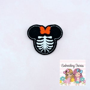May include: Black and white embroidered patch in the shape of a mouse head with a skeleton design. The mouse head has an orange bow and white stitching around the edges. The patch is on a white background.
