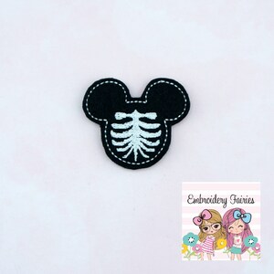 May include: A black felt applique in the shape of a mouse head with a light blue embroidered rib cage design. The applique has a white stitched border. The image also includes a logo for Embroidery Fairies.