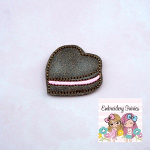 May include: A brown felt heart-shaped patch with a pink embroidered line across the centre. The patch has a glittery texture.