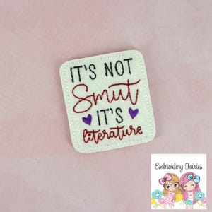 Op de afbeelding: Een rechthoekige patch van witte glitterstof met afgeronde hoeken, voorzien van zwart en rood geborduurd tekst die luidt IT'S NOT Smut IT'S LITERATURE, geaccentueerd door twee kleine paarse geborduurde hartjes tussen de tekstregels.