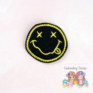 May include: Black felt embroidered patch with a yellow smiley face with crossed out eyes and a tongue sticking out. The patch is surrounded by a yellow stitched border.