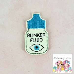 May include: A white and blue patch in the shape of a bottle of eye drops, with the words "BLINKER FLUID" and an eye graphic. The patch has a blue cap and a light blue eye with a black outline.