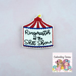 May include: Embroidered patch in the shape of a circus tent with the text "Ringmaster of the Shit Show". The tent is red and white with a blue and red border. A small yellow flag is at the top.