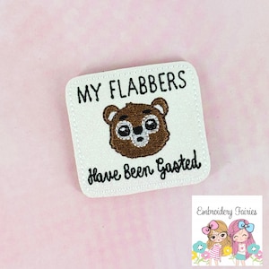 May include: Square white embroidered patch with the text "MY FLABBERS Have Been Gasted" and a brown bear face. The patch has a white stitched border. A small logo of two cartoon fairies is in the bottom right corner.