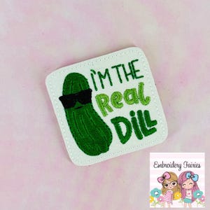 May include: Embroidered patch featuring a green pickle wearing black sunglasses. The text "I'M THE Real Dill" is embroidered in green. The patch is square with a white border and is on a pink background.
