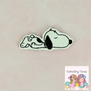 May include: A light green Snoopy patch with black outline and details, lying down with closed eyes. The patch has a glittery texture. The image also includes a logo for "Embroidery Fairies" with two cartoon girls.