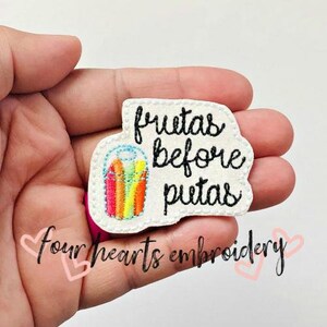 May include: A white embroidered patch with the text "frutas before putas" in black script. The patch features a colorful illustration of pencils in a cup. The words "four hearts embroidery" are also present.