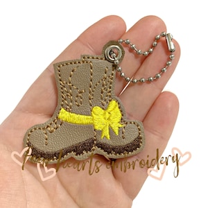 May include: A tan-colored boot-shaped keychain with yellow and brown embroidered details. The keychain features a yellow bow and decorative stitching. It is attached to a silver ball chain.